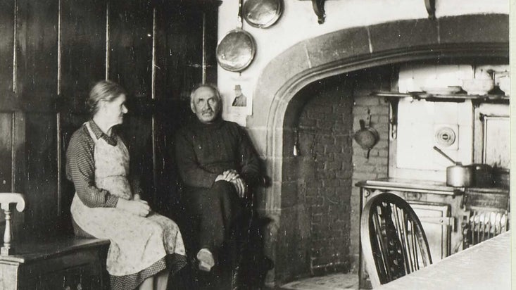 A black and white photograph of Mr and Mrs Lilburn sat in front of a large fireplace at Lindisfarne Castle with a dog walking in front of them